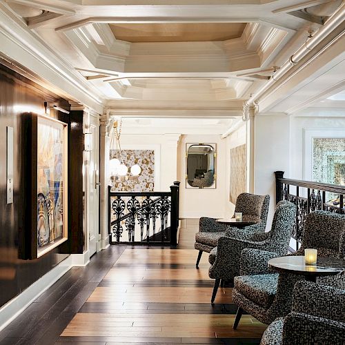 This image shows a stylish hallway with elegant seating, patterned armchairs, wall art, a mirror, wooden flooring, and intricate railing details.