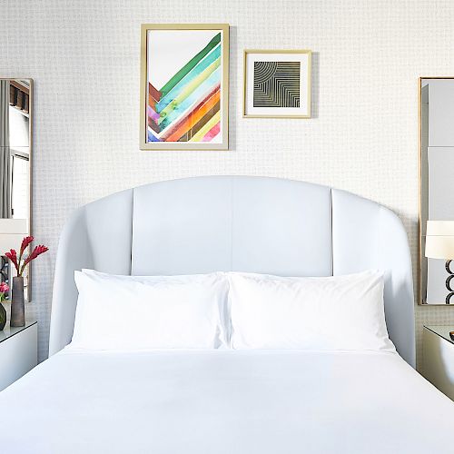 A neatly made bed with white linens, flanked by two nightstands holding identical lamps, with colorful artwork above the headboard.