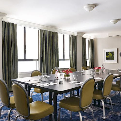 A modern conference room with a long table, surrounded by chairs and set for a meeting. There are windows with curtains and a TV on the wall.