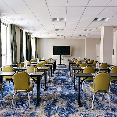 A room set up for a meeting or conference with rows of desks and chairs facing a large screen and windows along one side, carpeted floor.