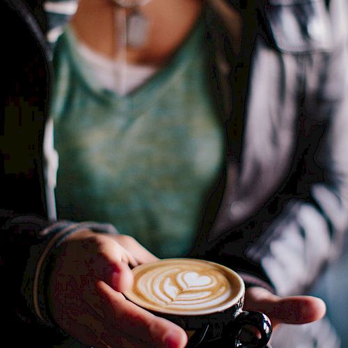 A person in a green shirt and leather jacket is holding a black cup of coffee with latte art. The scene has a warm and cozy ambiance.