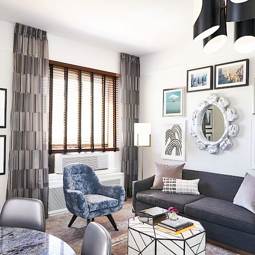 A modern living room with a grey sofa, blue armchair, patterned ottoman, wall art, circular mirror, window with blinds, and contemporary lighting.