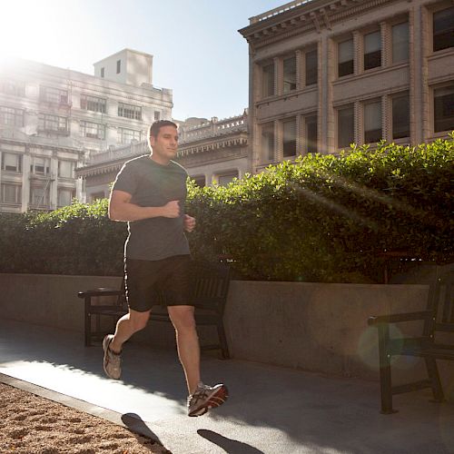 A person is jogging on a pathway in an urban area with tall buildings and greenery in the background during a sunny day.