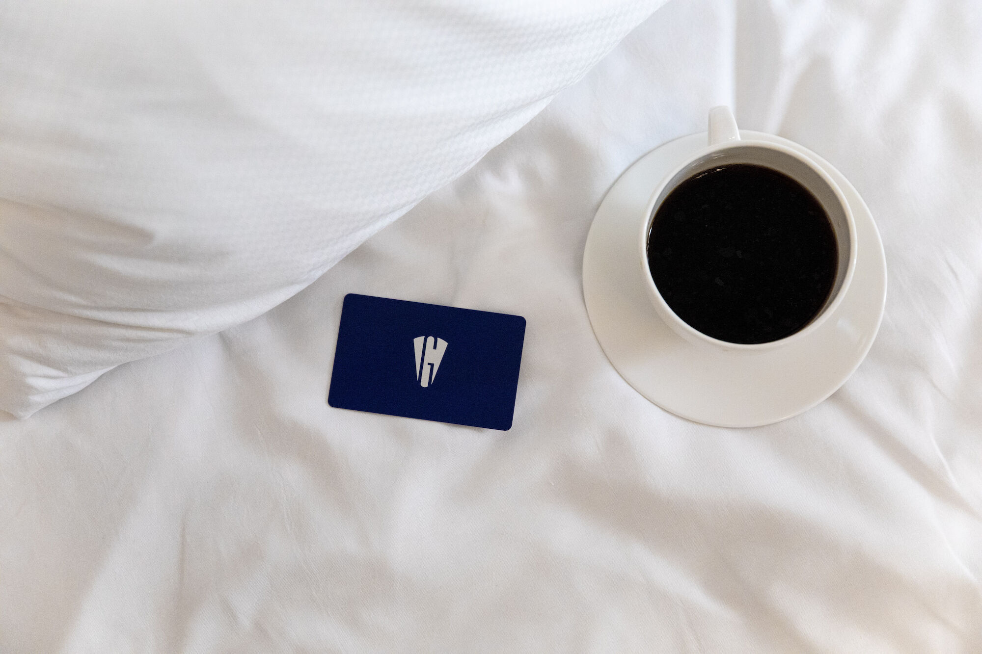 The image shows a cup of coffee and a blue card on a white bed with a pillow.