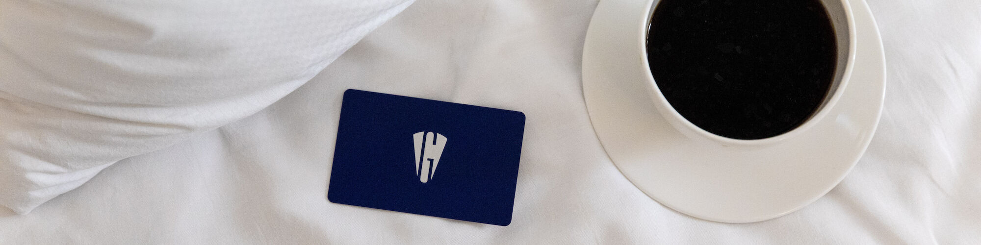 The image shows a cup of coffee and a blue card on a white bed with a pillow.