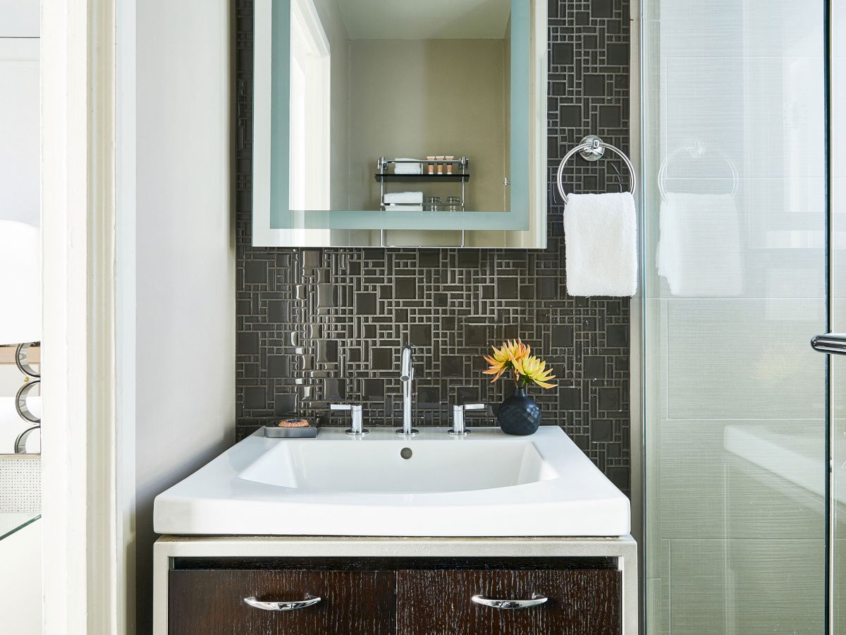 A modern bathroom sink with a mirror, towel ring, and a flower vase beside it. There's a glass shower door to the right, finishing the sentence.