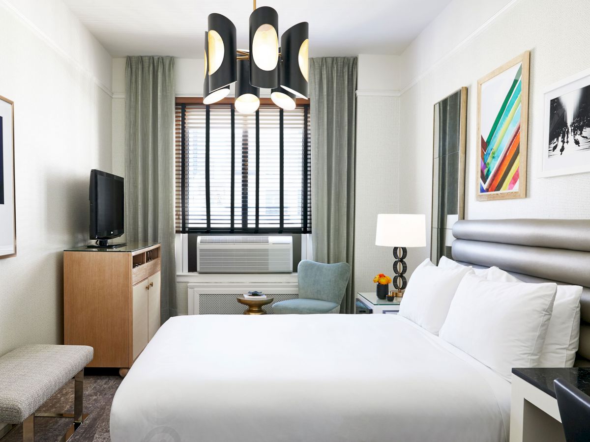 This image shows a modern hotel room with a bed, artwork on the walls, a TV, a small table, a chair, an air conditioner, and a unique light fixture overhead.