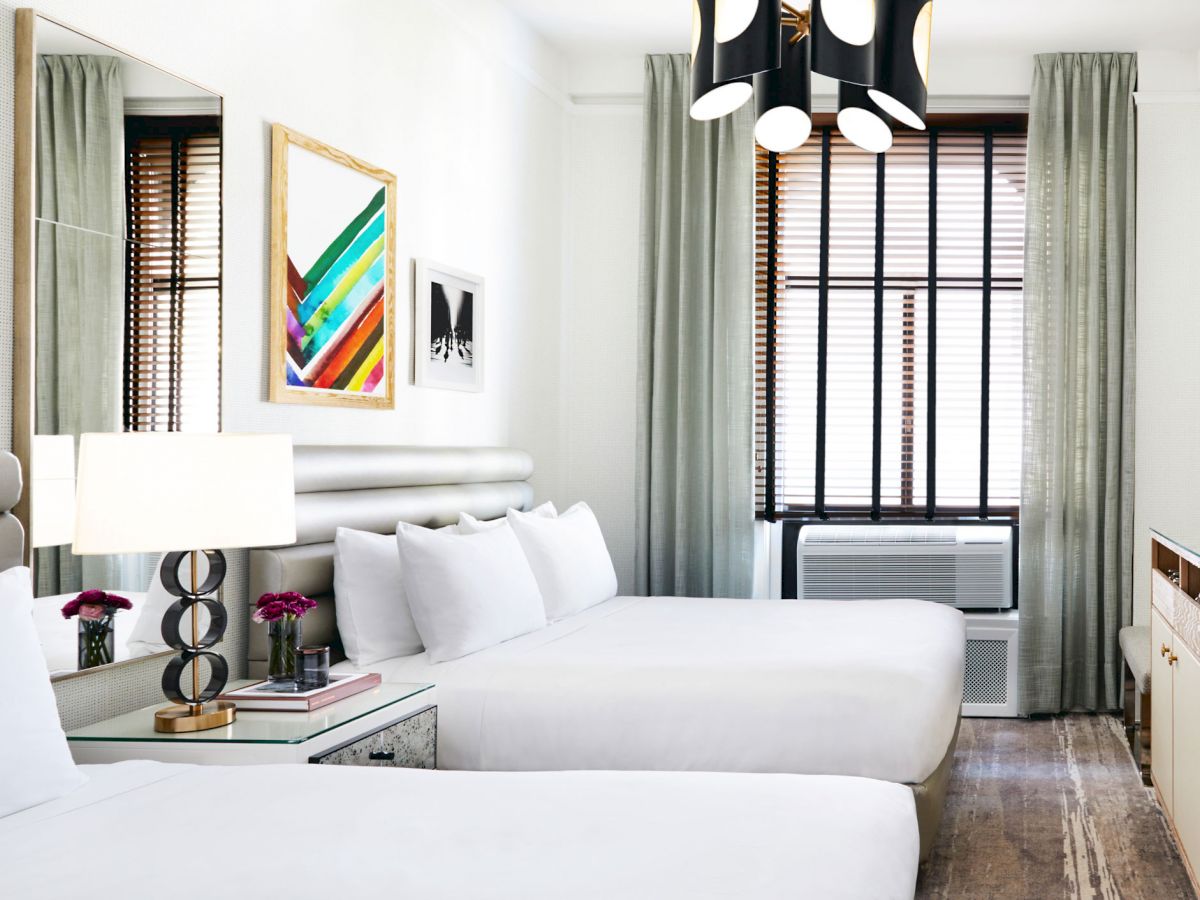 The image shows a modern hotel room with two beds, a TV, colorful artwork, and a bedside lamp, featuring minimalist decor and natural light.