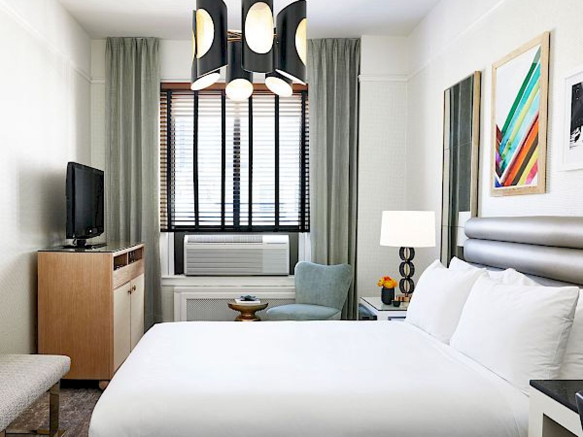 A modern hotel room with a white bed, stylish lighting, a TV on a wooden cabinet, and an air conditioner under the window with curtains.