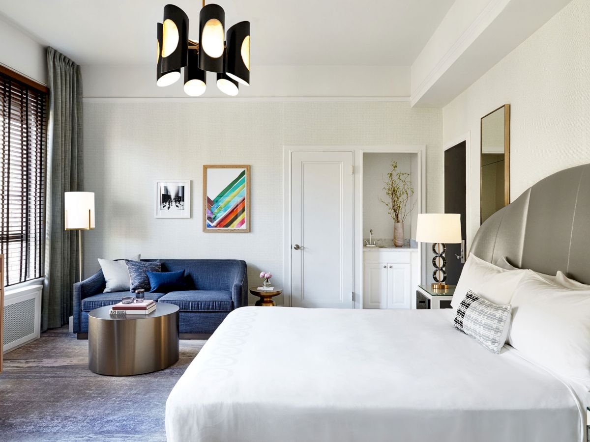 A modern bedroom with a bed, a blue sofa, framed art, and various furnishings, all illuminated by natural light and stylish ceiling lights.