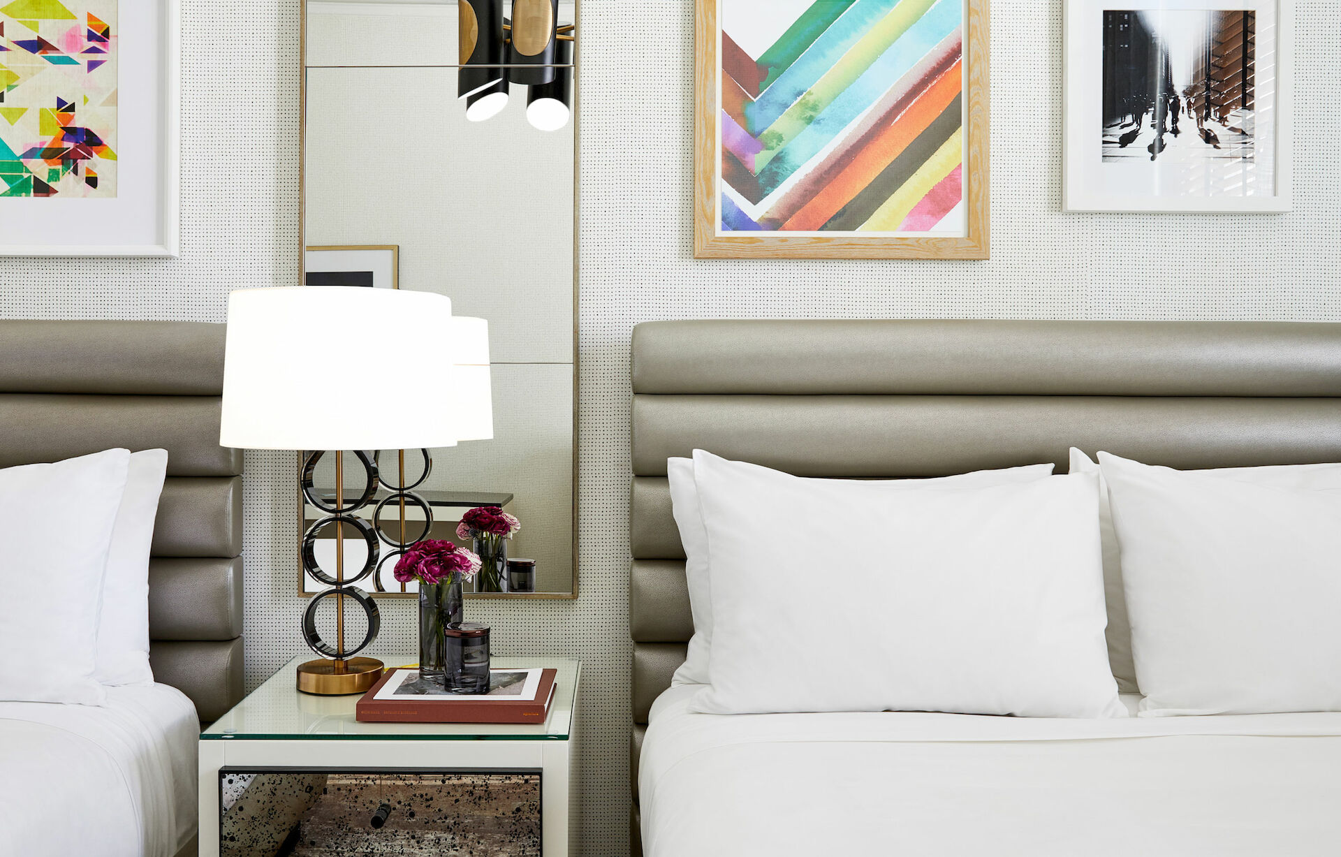 The image shows a modern hotel room with two beds, a lamp on a nightstand, and colorful artwork on the wall above the beds.