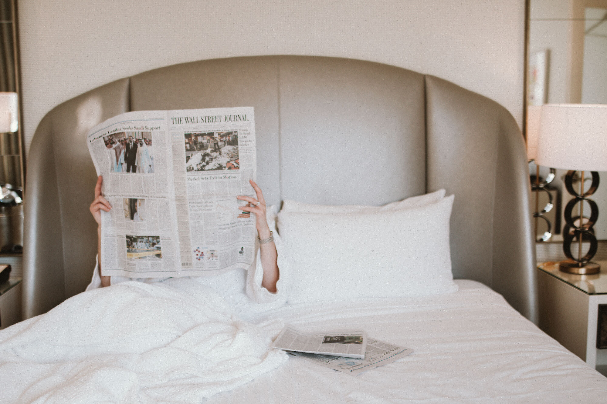 A person is sitting in bed, reading a newspaper, with a lamp and a smaller newspaper on the bed.