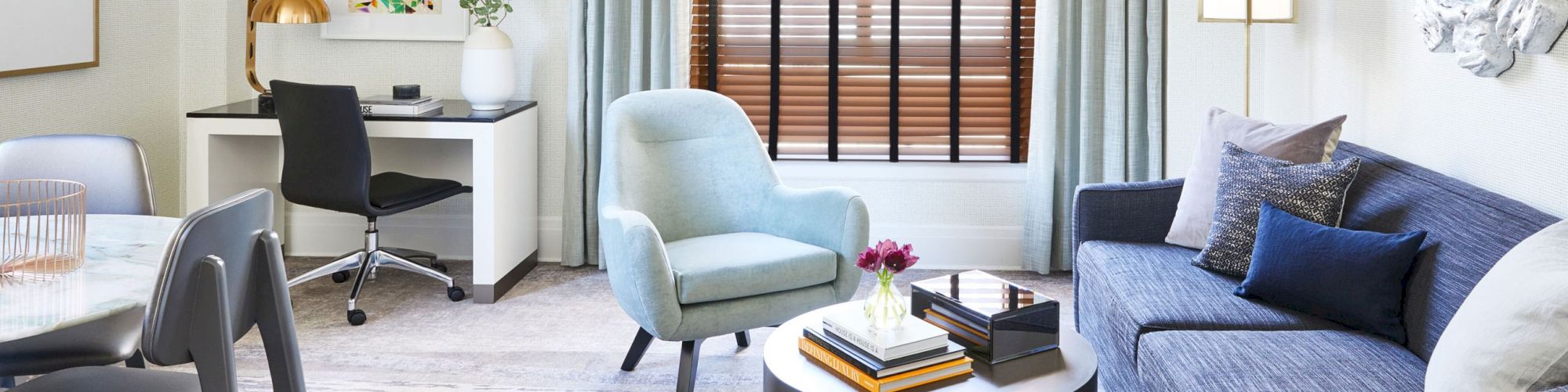 A modern living room with a blue armchair, gray sofa, coffee table, desk, and decor. There's a window with blinds and curtains.