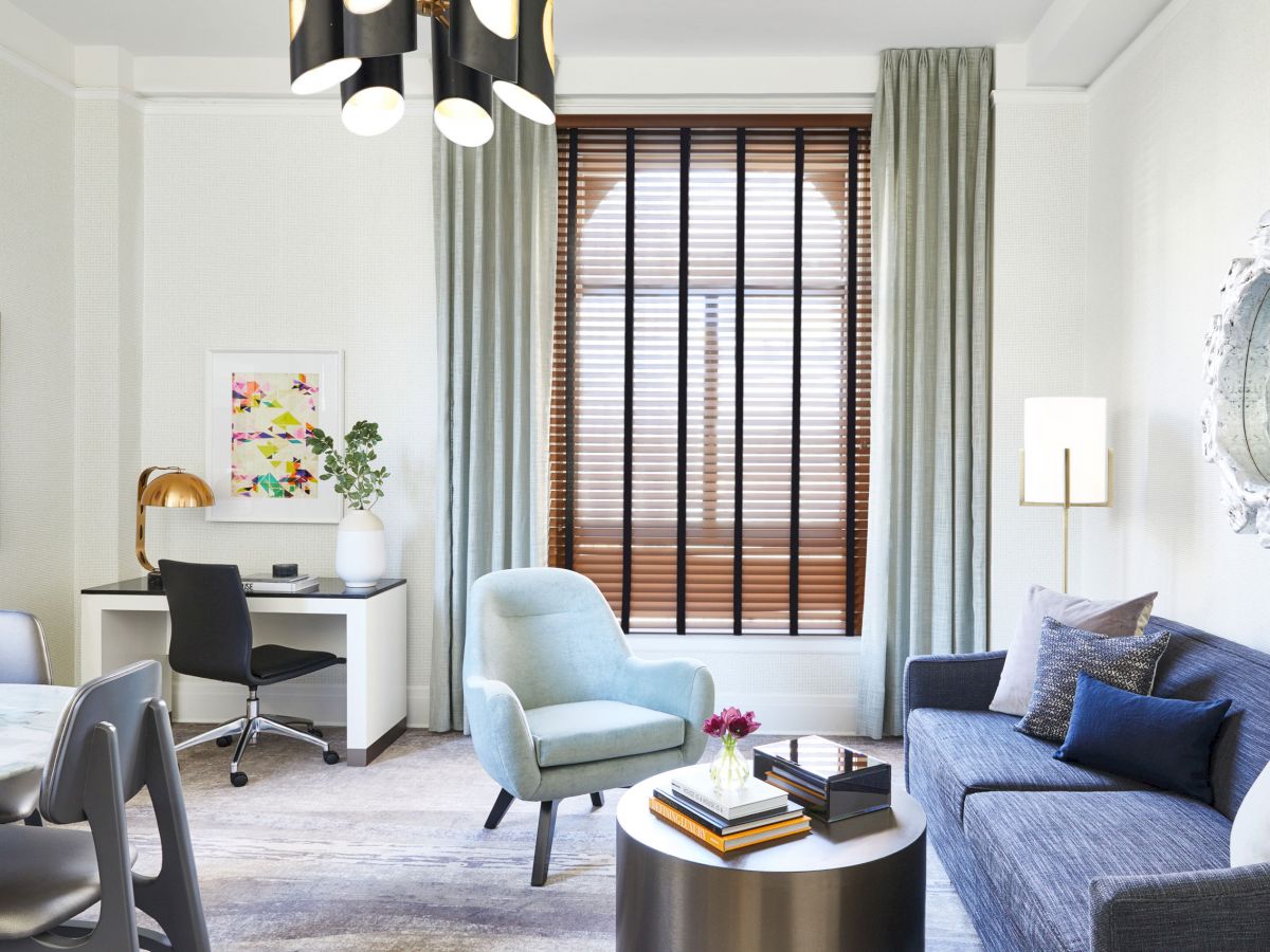 A modern living room with a blue couch, armchair, coffee table, desk, and chair. Large window with blinds, wall art, and chic decor complete the space.