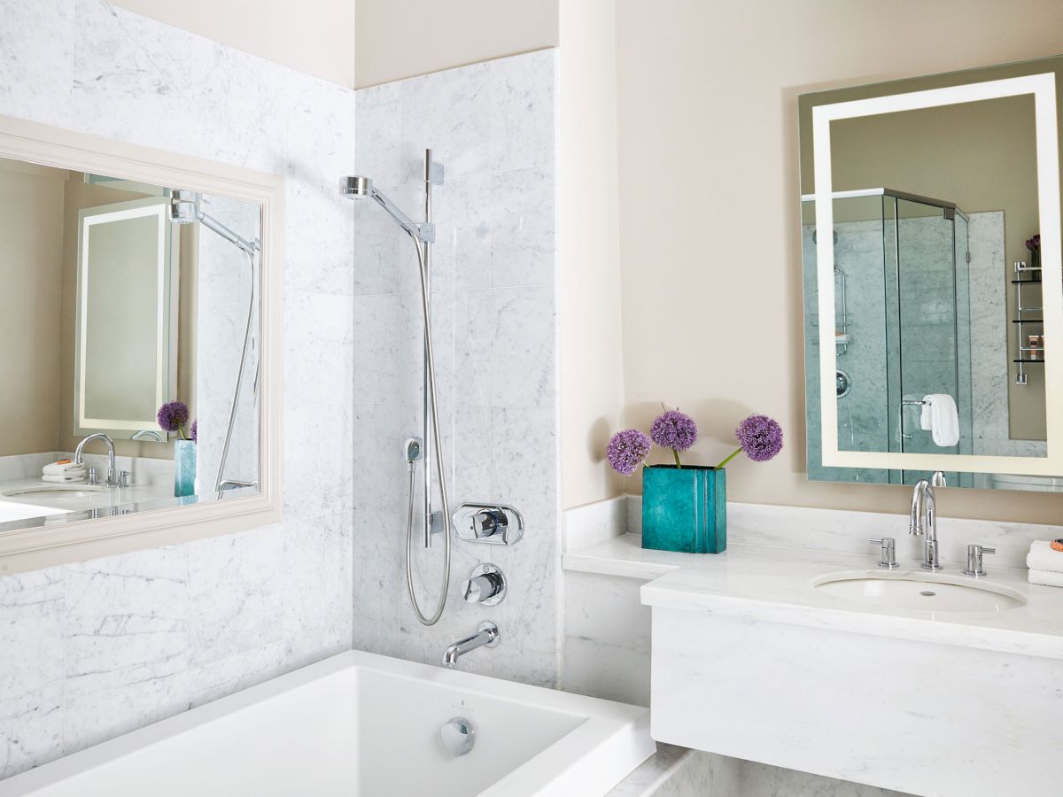 A modern bathroom features a bathtub, showerhead, mirror, sink, and vase with purple flowers on the counter, bathed in natural light.