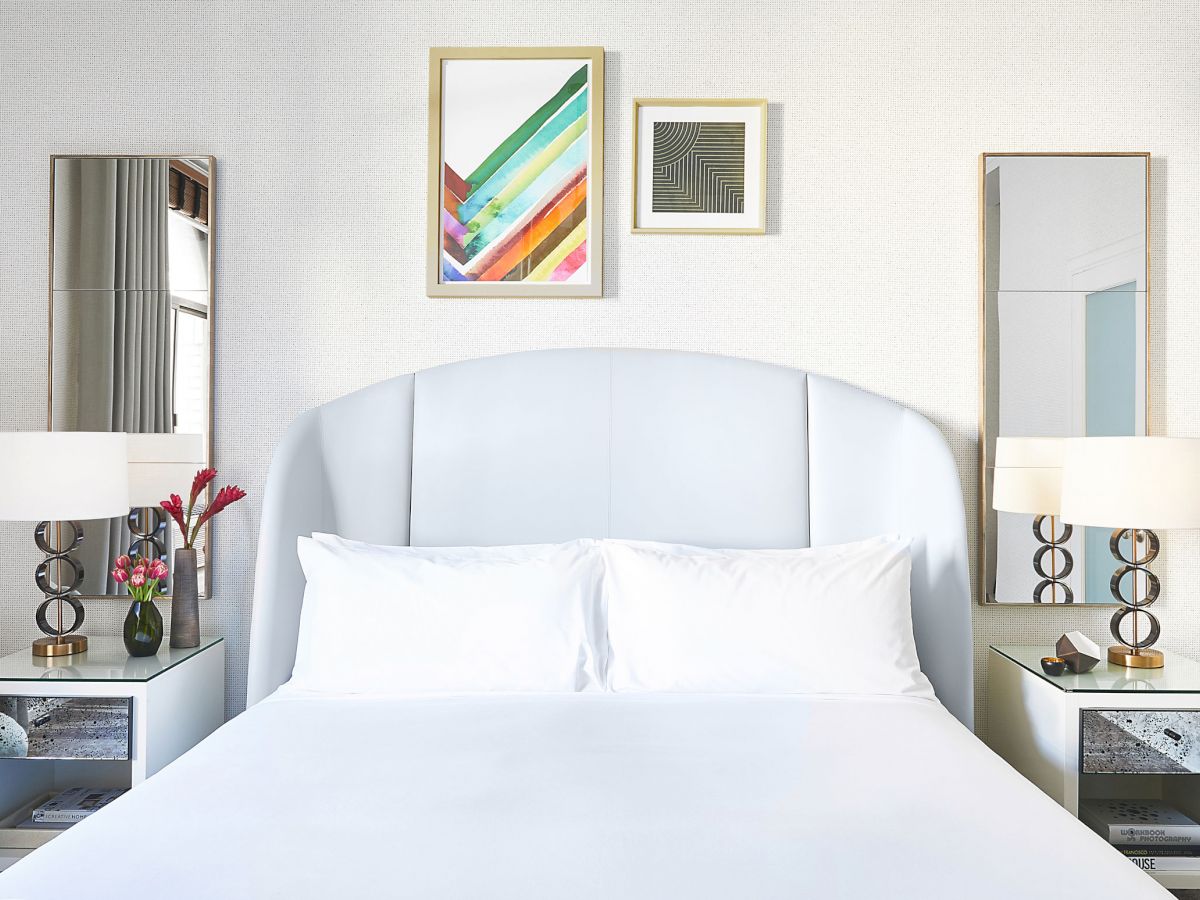 A neatly arranged bedroom with white bedding, two side tables with lamps, mirrored wall decor, and colorful abstract art prints above the bed.