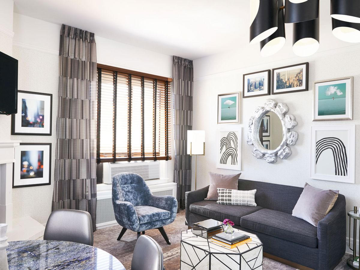 A modern living room with a gray couch, blue armchair, geometric coffee table, wall art, and a mirror. Natural light flows in from the window.