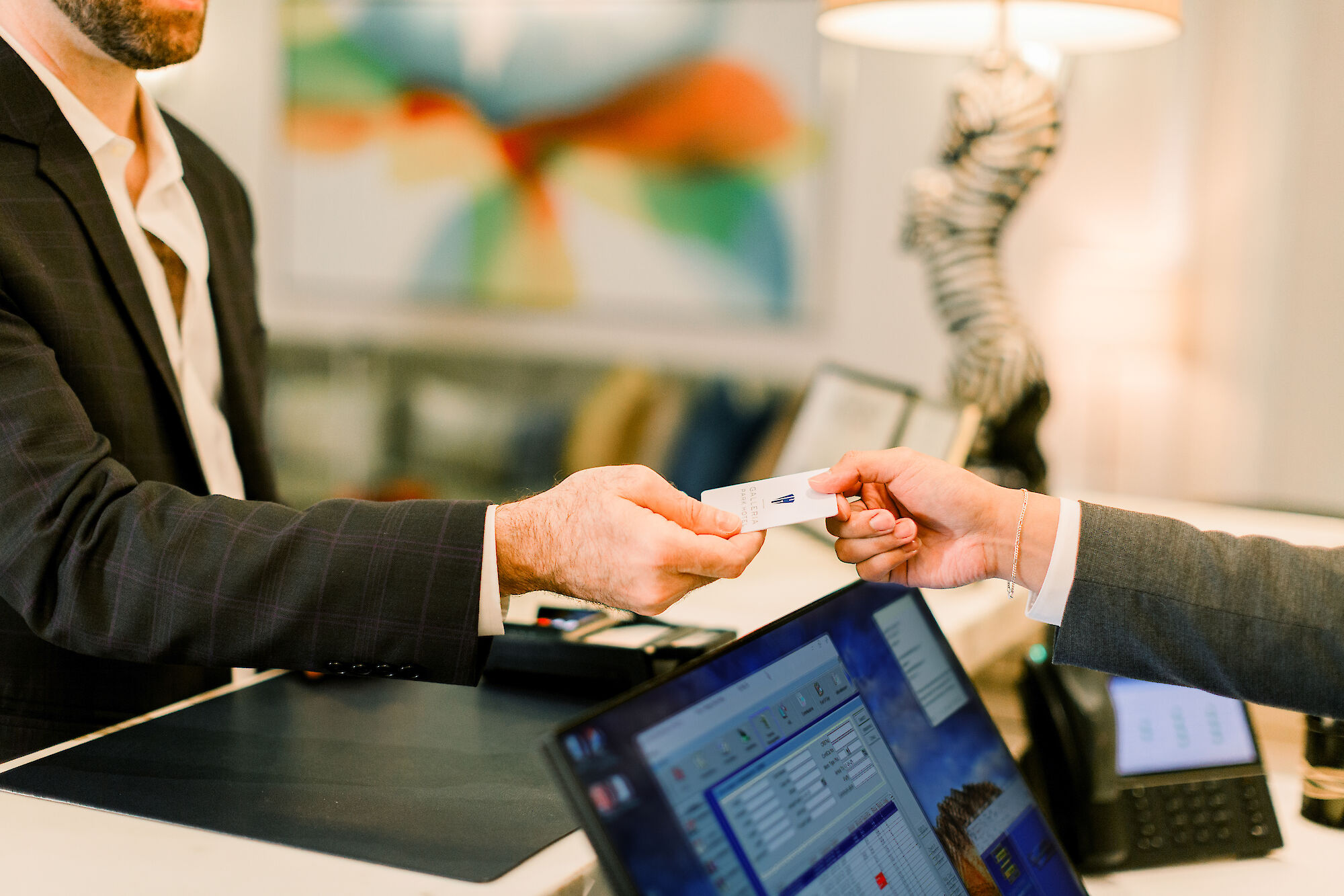 Two people are exchanging a key card at a reception desk, with a computer visible in the foreground.