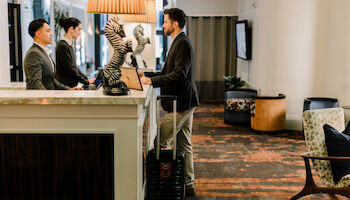 A man with a suitcase stands at a hotel reception desk, talking to two staff members. The desk features zebra-themed lamps.