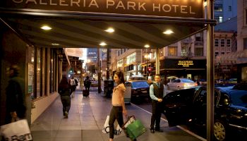 People are walking with shopping bags under the illuminated sign of Galleria Park Hotel in a busy urban street at dusk.