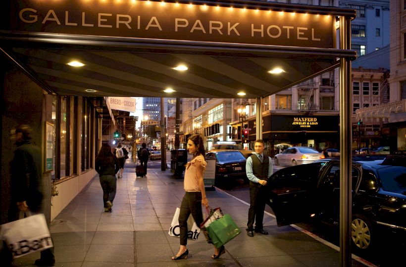 People are walking with shopping bags under the illuminated sign of Galleria Park Hotel in a busy urban street at dusk.