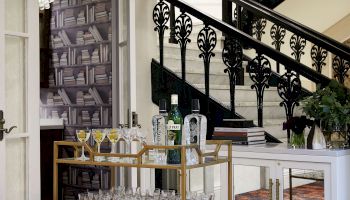 A stylish home bar cart with glasses and liquor bottles in front of a staircase with ornate railing, next to a white cabinet and a book-patterned wall.