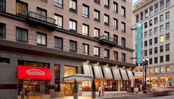 The image shows a city street, with a building featuring shops on the ground floor, including a red awning restaurant, and a hotel sign.