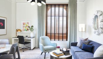 A modern living room with a desk, chair, armchair, sofa, coffee table, floor lamp, wall art, and large window with blinds and curtains.