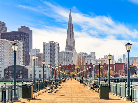 A boardwalk leading to a city skyline with a pyramid-shaped building, surrounded by skyscrapers and street lamps under a blue sky.