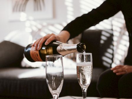 A person pouring champagne or sparkling wine into two glasses on a table with a cork nearby and modern decor in the background.