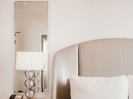 A neatly made bed with white linens, a bedside table with a lamp, clock, and phone, and a wall mirror in a minimalist bedroom.