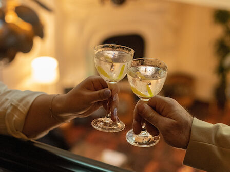 Two people clinking glasses in a warmly lit room, viewed from the side above a balcony railing.