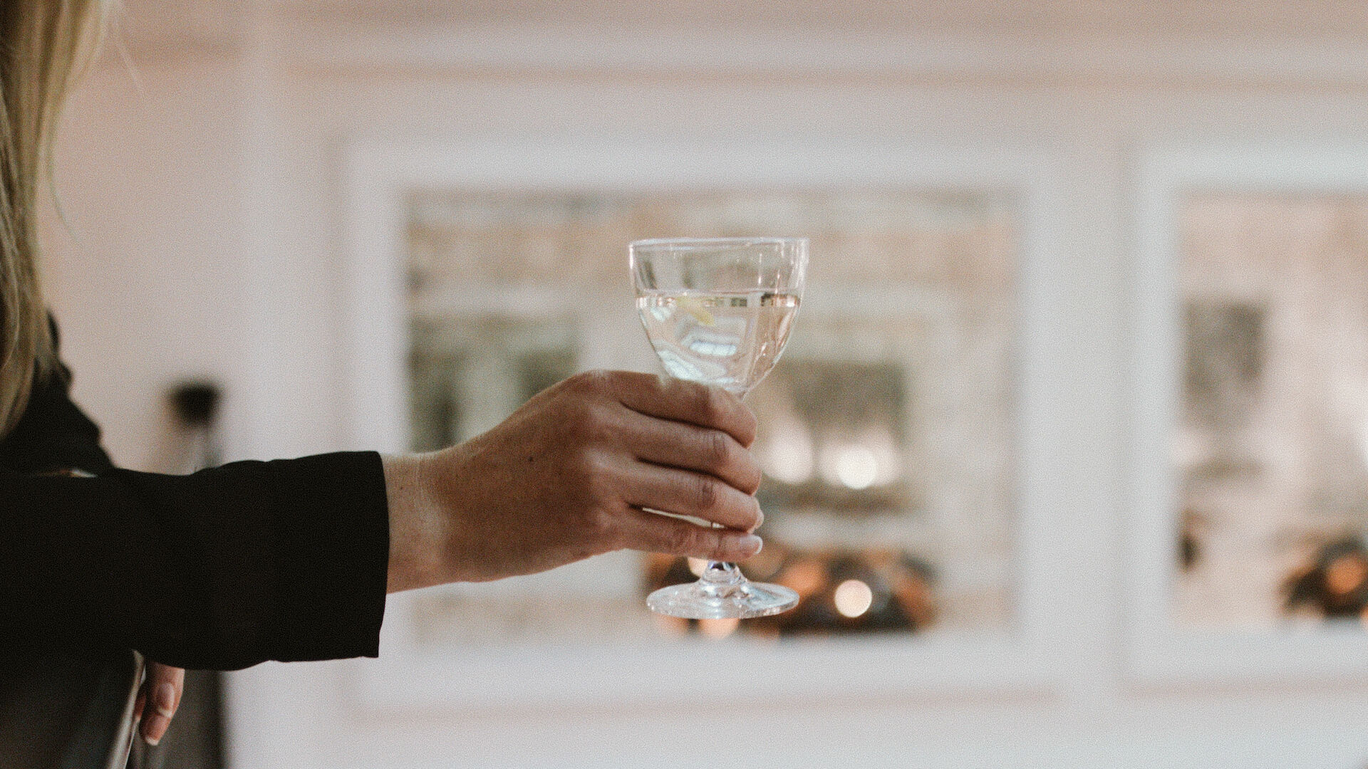 A person leaning on a balcony holding a wine glass, indoor fancy setting with blurred lights in the background.