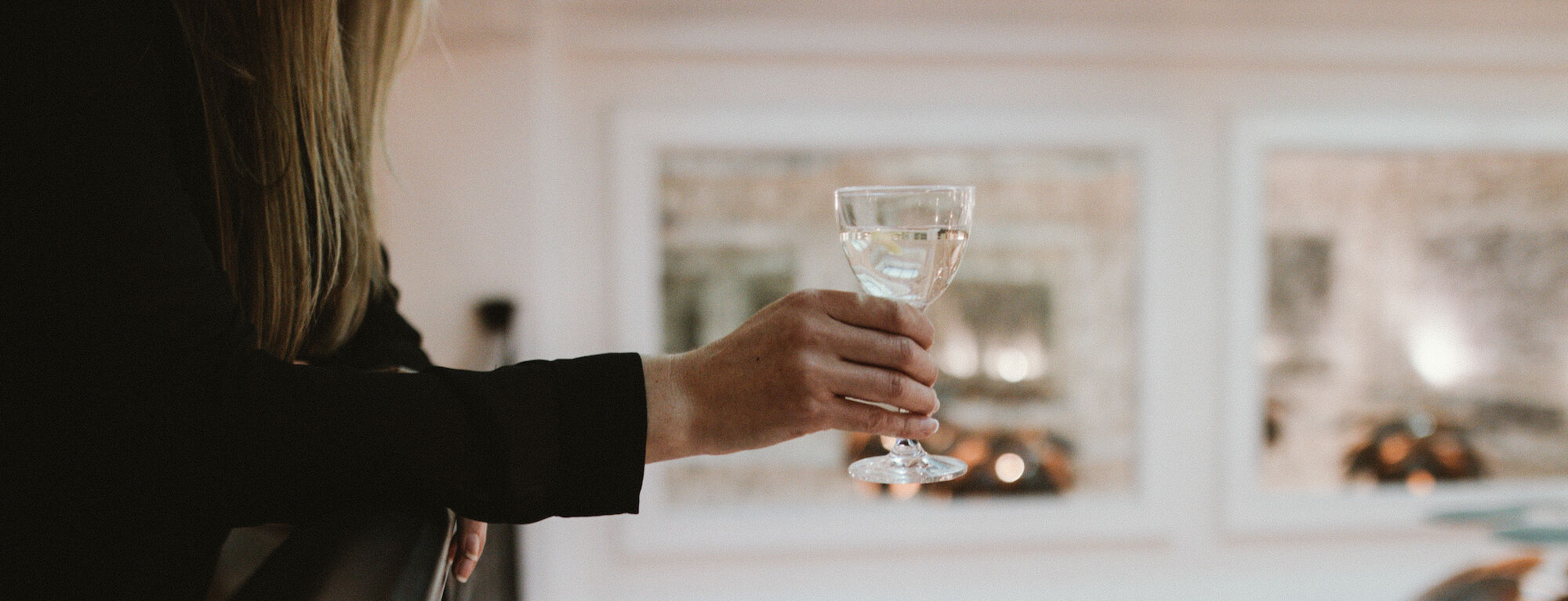 A person leaning on a balcony holding a wine glass, indoor fancy setting with blurred lights in the background.