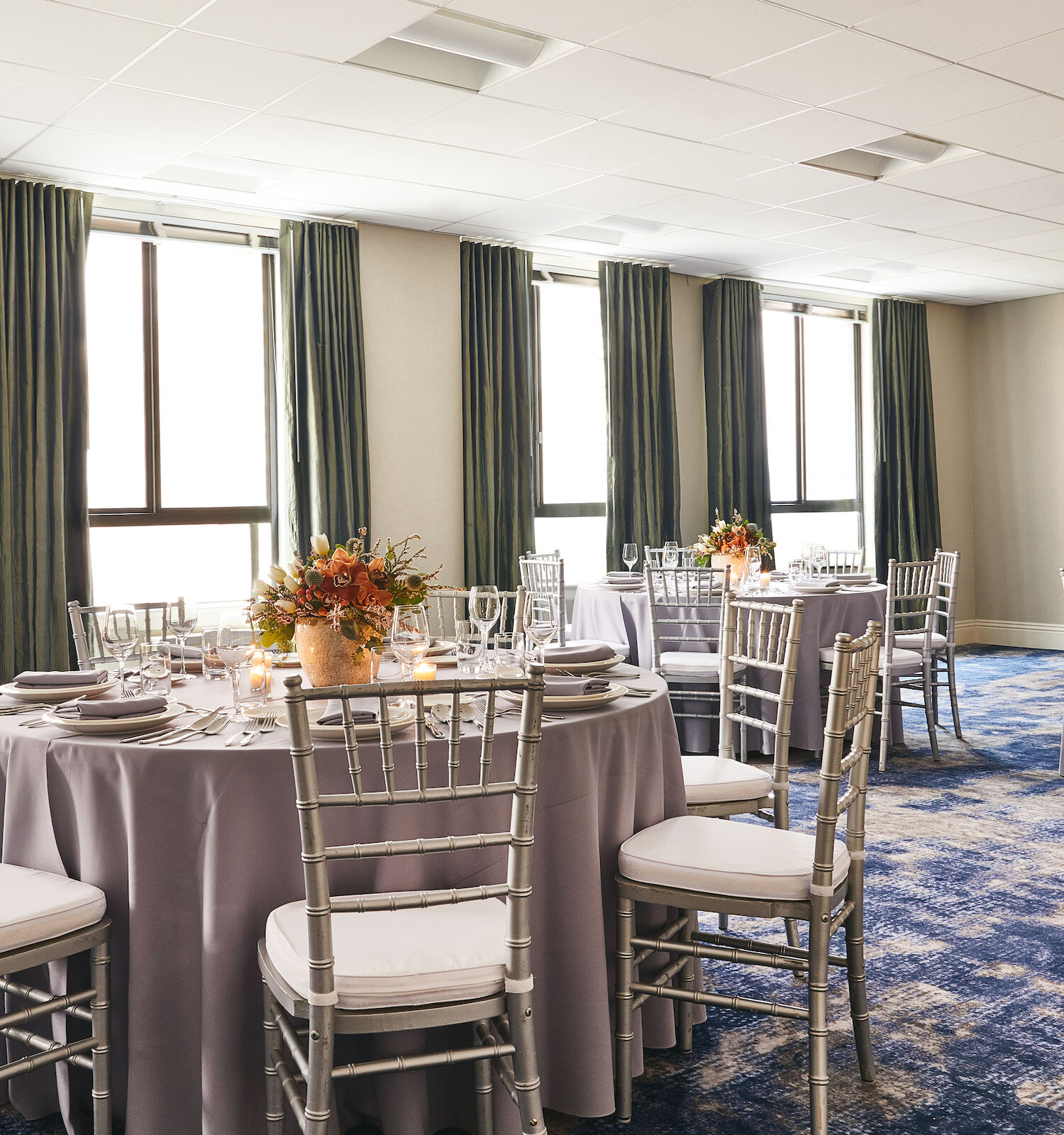 A formal banquet room with round tables set with glassware, floral centerpieces, candles, and neatly arranged chairs by tall windows.