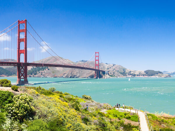 Golden Gate Bridge spanning turquoise bay, with green hills in foreground, sailboats in the water, and a clear blue sky above.