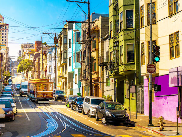 A vibrant San Francisco&ndash;style street with colorful houses, parked cars, trolley tracks, and sunny skies, lively neighborhood vibes.