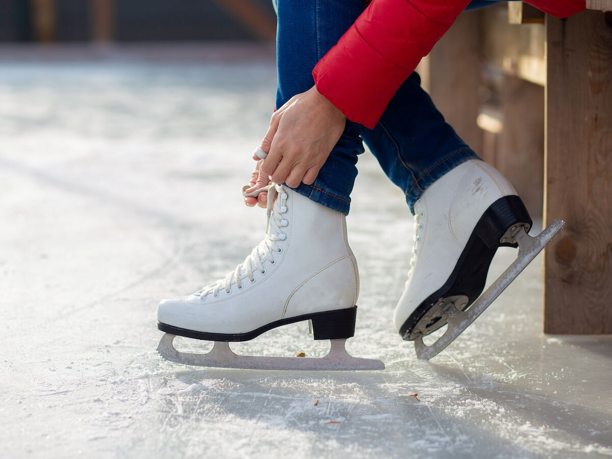 A person wearing a red jacket laces up white ice skates while sitting on a bench on the ice rink.