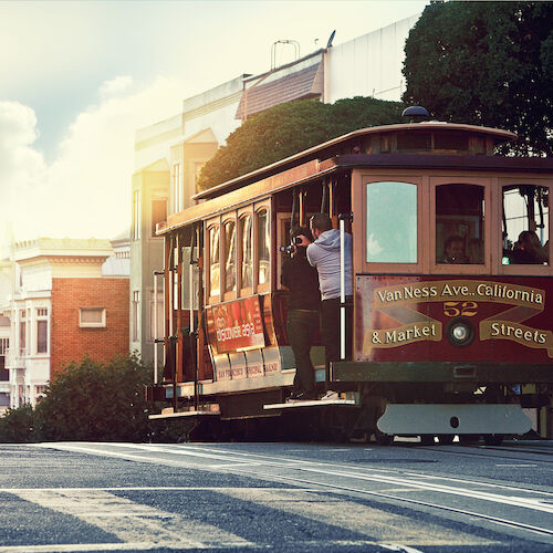 A classic San Francisco cable car travels along a street, with picturesque buildings and trees in the background, capturing evening light.