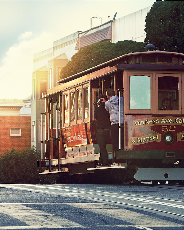 A classic San Francisco cable car travels along a street, with picturesque buildings and trees in the background, capturing evening light.