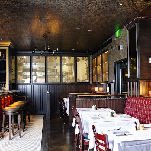 The image shows a stylish restaurant interior with a bar, red cushioned seating, tables set with white cloth, and elegant dark wood decor.