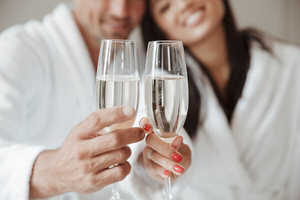 A couple in white robes clinks champagne glasses, smiling and celebrating together.