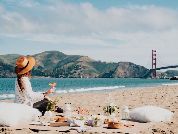 A woman in a sun hat has a seaside picnic on a sandy beach near the Golden Gate Bridge backdrop, with a cozy setup and fluffy pillows.