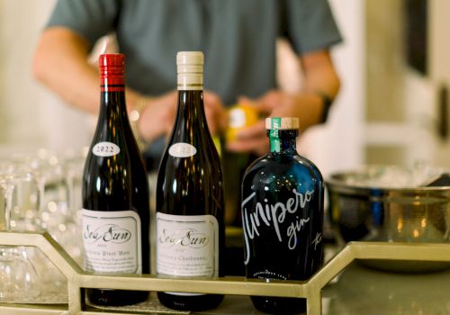 The image shows two bottles of wine and a bottle of gin on a bar counter, with a person preparing drinks in the background.