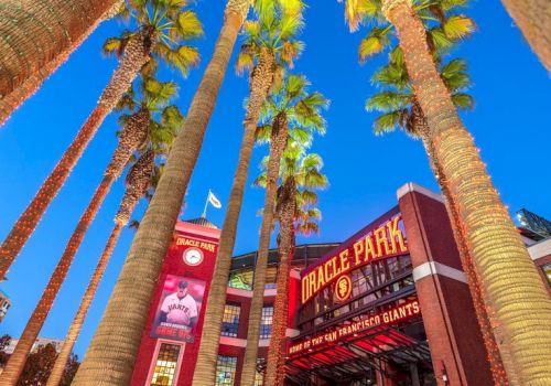 The image shows a view looking up at tall palm trees with the entrance to Oracle Park, a baseball stadium, in the background under a clear blue sky.