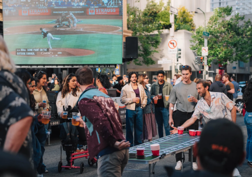 A group of people enjoys a street gathering with a game, drinks, and a large screen showing a baseball game in the background.