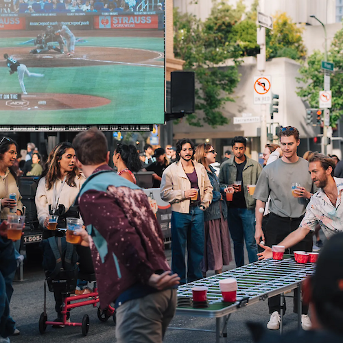 A group of people enjoys a street gathering with a game, drinks, and a large screen showing a baseball game in the background.