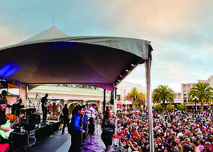 A band is performing on a stage under a tent, with a large crowd gathered to watch in an outdoor setting with palm trees.