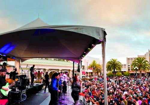 A band is performing on a stage under a tent, with a large crowd gathered to watch in an outdoor setting with palm trees.
