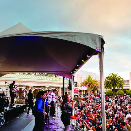 A band is performing on a stage under a tent, with a large crowd gathered to watch in an outdoor setting with palm trees.